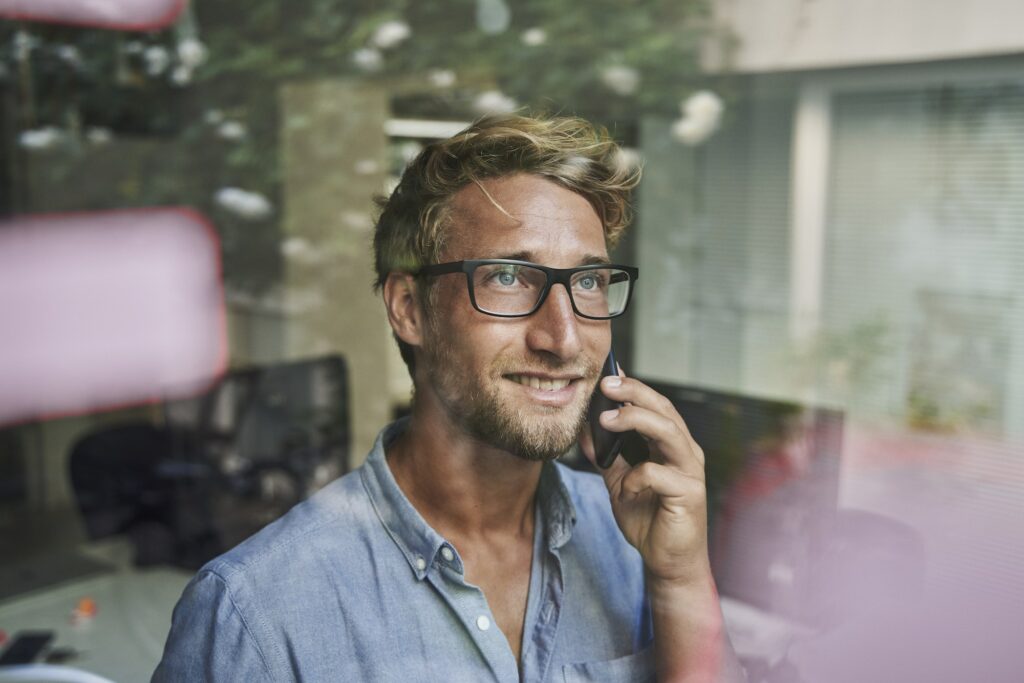 Casual young businessman on the phone behind windowpane in office